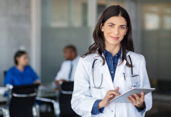 Portrait of beautiful mature woman doctor holding digital tablet and looking at camera. Confident female doctor using digital tablet with colleague talking in background at hospital. Latin nurse in labcoat and stethoscope in private clinic with medical team working.