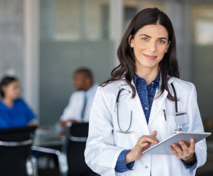 Portrait of beautiful mature woman doctor holding digital tablet and looking at camera. Confident female doctor using digital tablet with colleague talking in background at hospital. Latin nurse in labcoat and stethoscope in private clinic with medical team working.